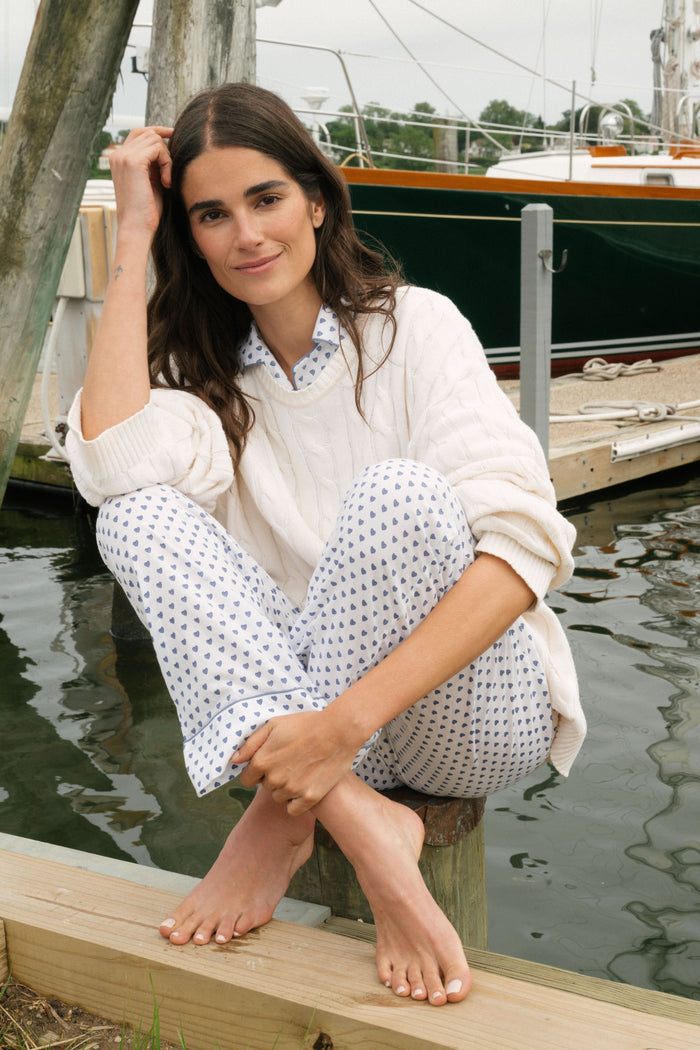A person stands barefoot in Roller Rabbit’s Hearts Loungewear, a matching cotton pajama set with heart prints, featuring a button-up long sleeve top and long pants. The background is plain white.