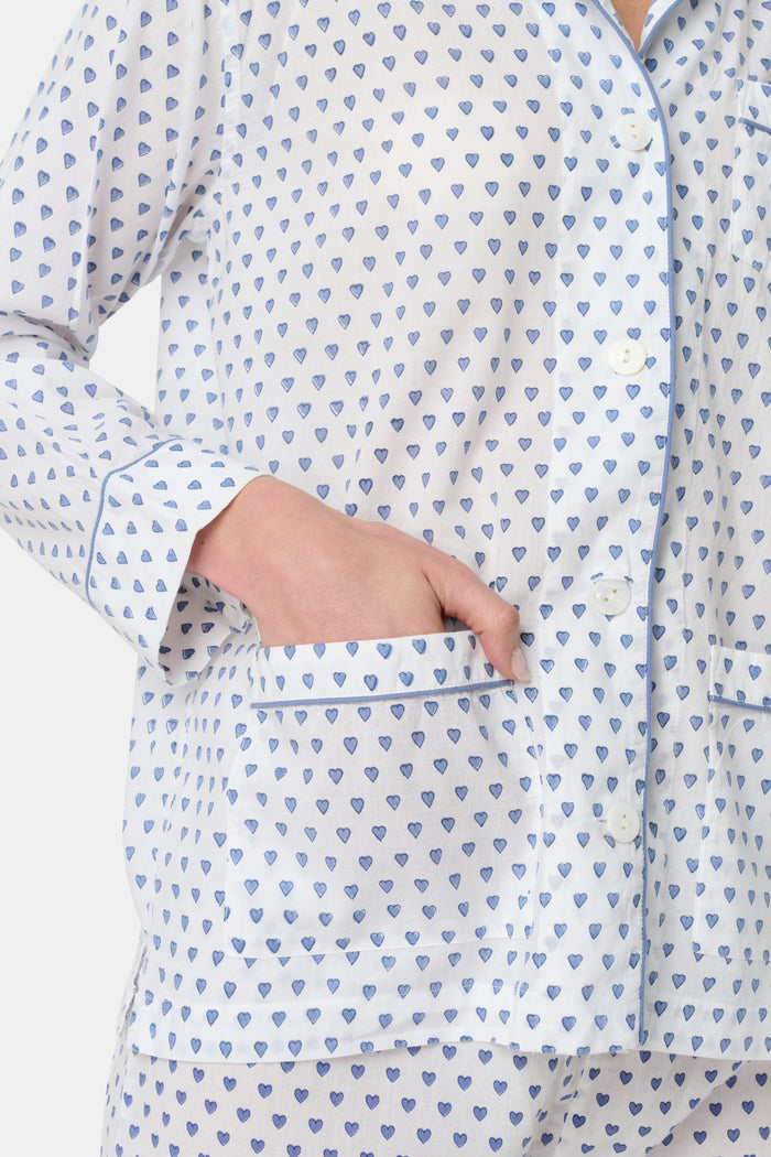 A person stands barefoot in Roller Rabbit’s Hearts Loungewear, a matching cotton pajama set with heart prints, featuring a button-up long sleeve top and long pants. The background is plain white.