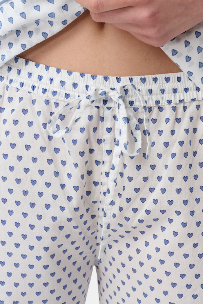 A person stands barefoot in Roller Rabbit’s Hearts Loungewear, a matching cotton pajama set with heart prints, featuring a button-up long sleeve top and long pants. The background is plain white.