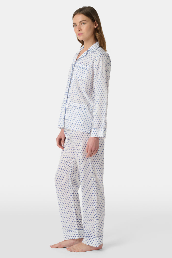A person stands barefoot in Roller Rabbit’s Hearts Loungewear, a matching cotton pajama set with heart prints, featuring a button-up long sleeve top and long pants. The background is plain white.