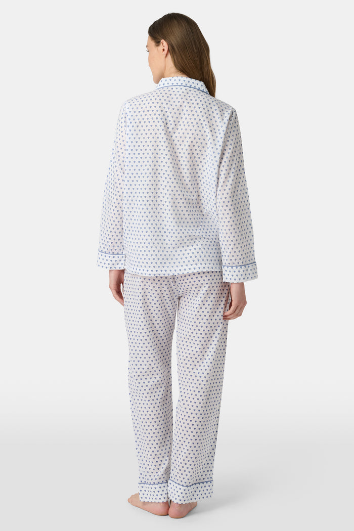A person stands barefoot in Roller Rabbit’s Hearts Loungewear, a matching cotton pajama set with heart prints, featuring a button-up long sleeve top and long pants. The background is plain white.