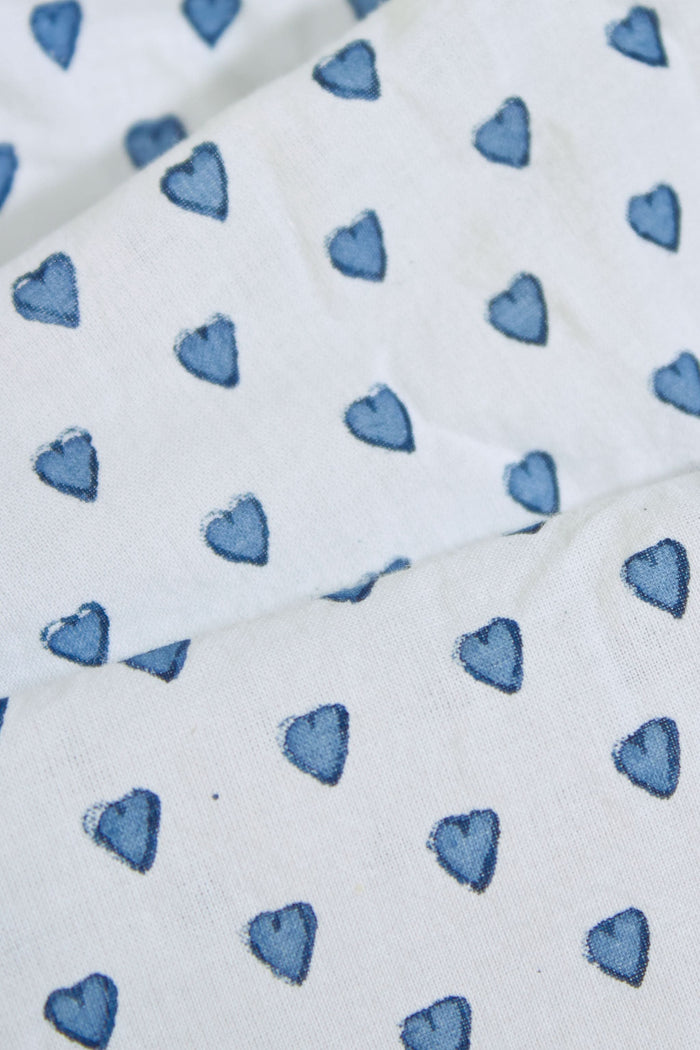A person stands barefoot in Roller Rabbit’s Hearts Loungewear, a matching cotton pajama set with heart prints, featuring a button-up long sleeve top and long pants. The background is plain white.