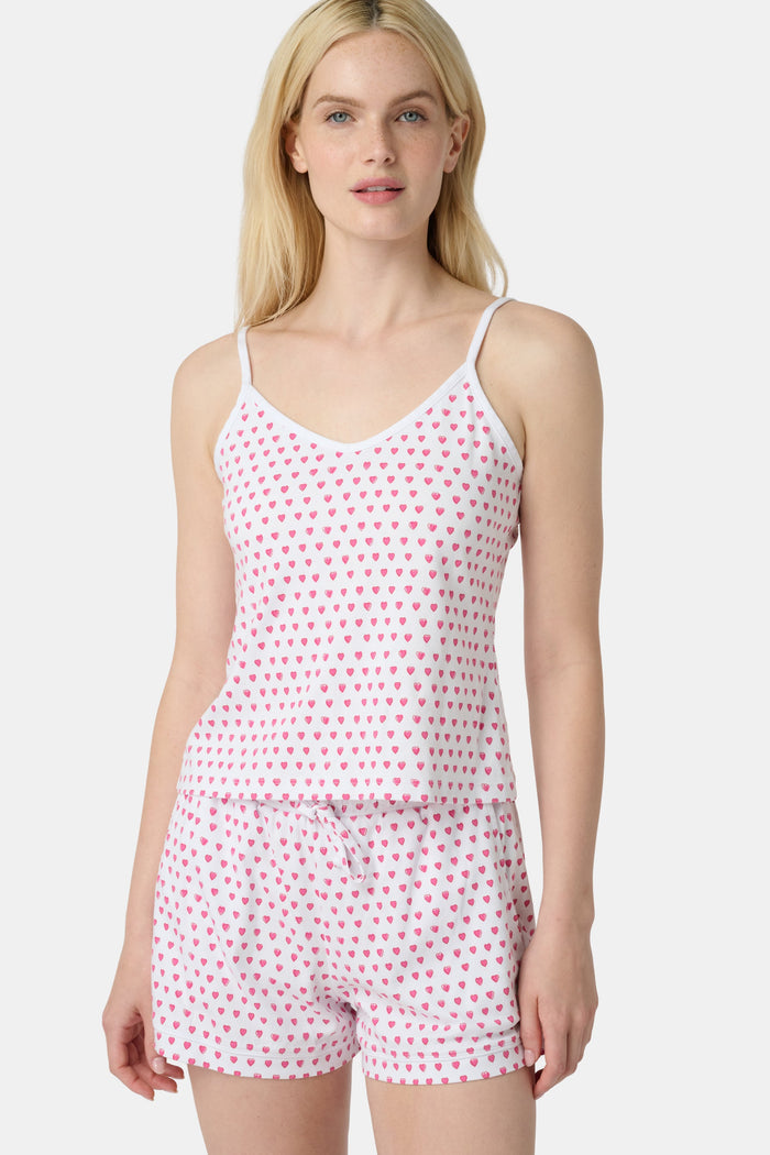 A woman wears the Roller Rabbit Hearts Mischa Set, a matching white loungewear set with pink hearts, featuring a sleeveless camisole and shorts made from soft Pima Cotton, standing against a plain white background.