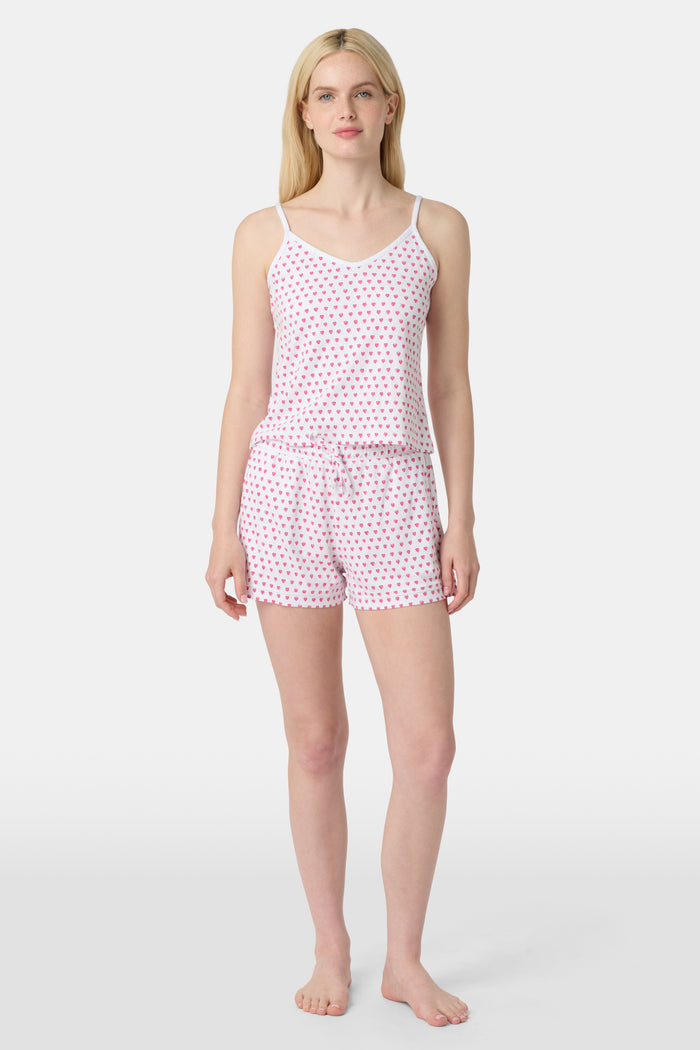 A woman wears the Roller Rabbit Hearts Mischa Set, a matching white loungewear set with pink hearts, featuring a sleeveless camisole and shorts made from soft Pima Cotton, standing against a plain white background.