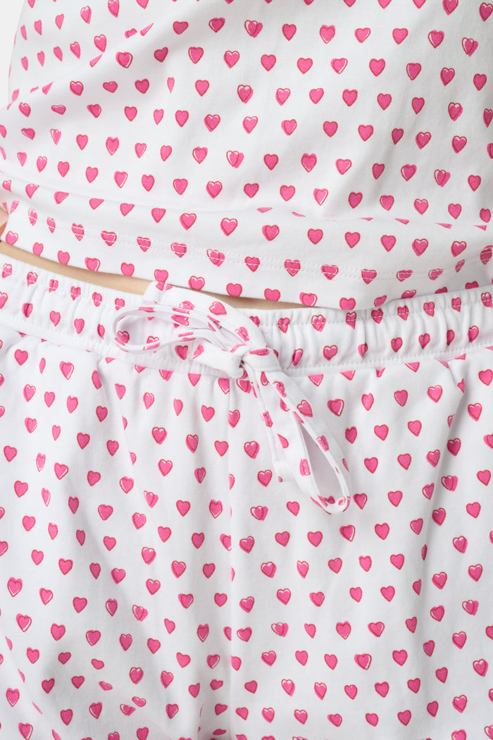 A woman wears the Roller Rabbit Hearts Mischa Set, a matching white loungewear set with pink hearts, featuring a sleeveless camisole and shorts made from soft Pima Cotton, standing against a plain white background.