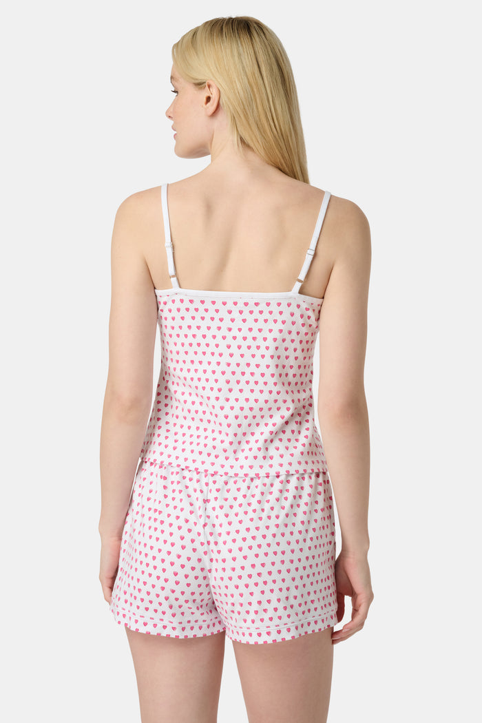 A woman wears the Roller Rabbit Hearts Mischa Set, a matching white loungewear set with pink hearts, featuring a sleeveless camisole and shorts made from soft Pima Cotton, standing against a plain white background.