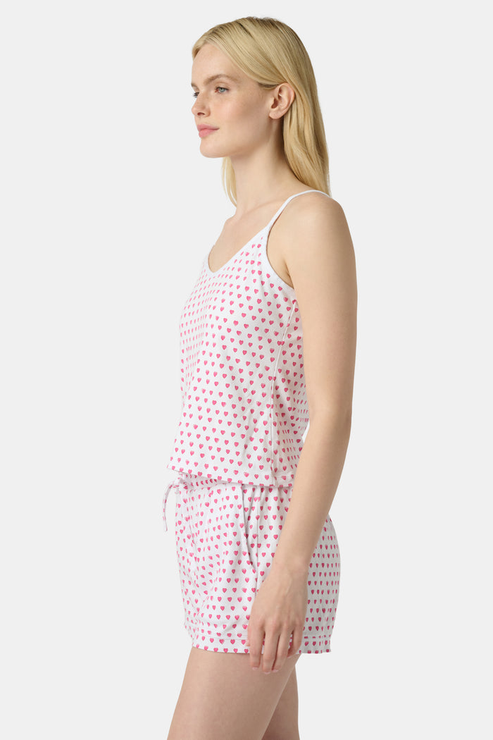 A woman wears the Roller Rabbit Hearts Mischa Set, a matching white loungewear set with pink hearts, featuring a sleeveless camisole and shorts made from soft Pima Cotton, standing against a plain white background.