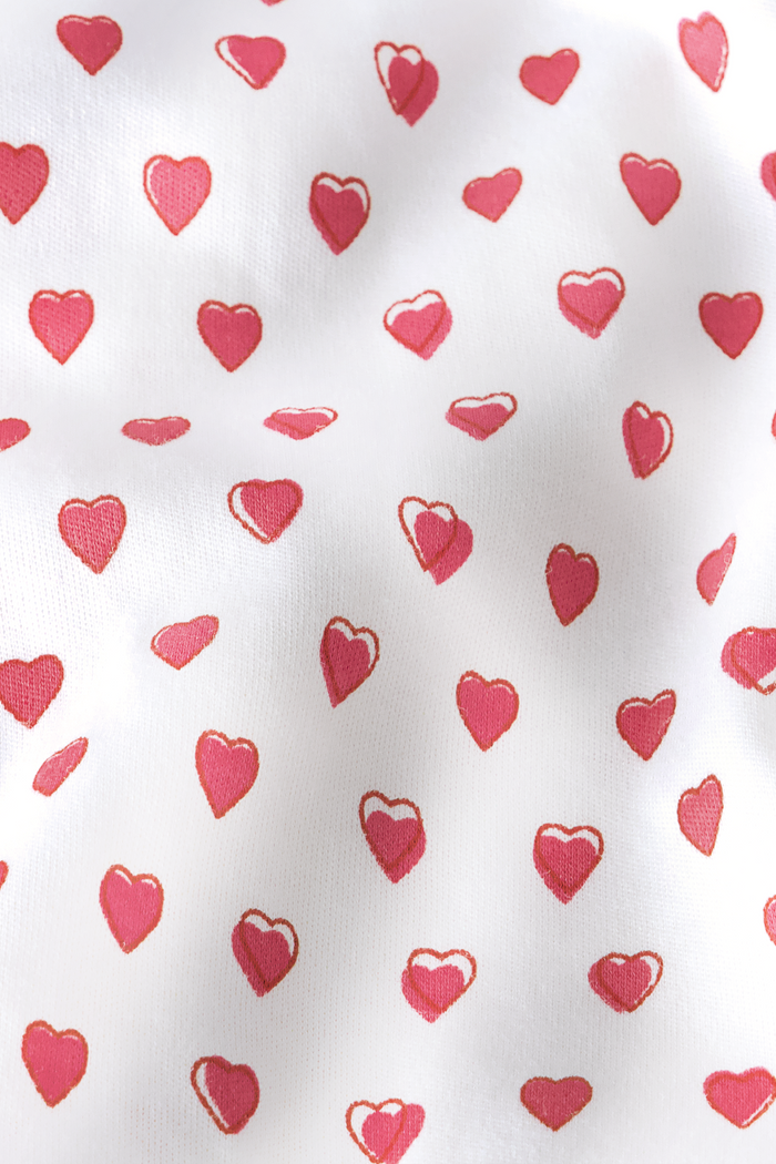 A person stands barefoot in Roller Rabbit’s Hearts Fiora Pajamas—light pink with red hearts. The matching set features a short-sleeve button-up top and long pants, made from soft Pima cotton, against a plain white background.