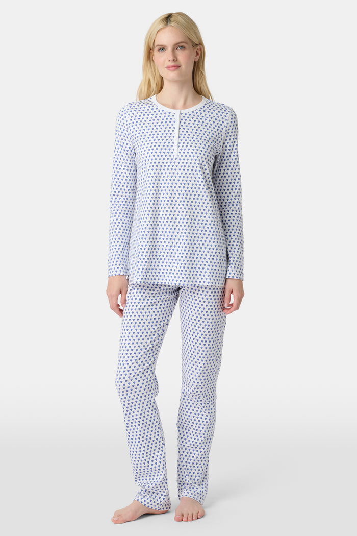A person with light skin and long blonde hair stands barefoot in Roller Rabbit’s Hearts Pajamas—a matching set made from soft Pima cotton, featuring a white background with small blue and red heart patterns.