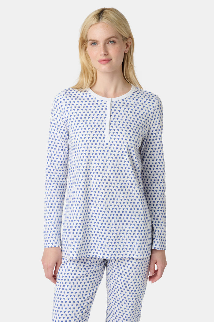 A person with light skin and long blonde hair stands barefoot in Roller Rabbit’s Hearts Pajamas—a matching set made from soft Pima cotton, featuring a white background with small blue and red heart patterns.