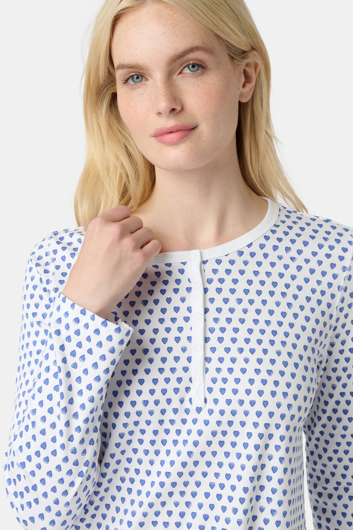 A person with light skin and long blonde hair stands barefoot in Roller Rabbit’s Hearts Pajamas—a matching set made from soft Pima cotton, featuring a white background with small blue and red heart patterns.