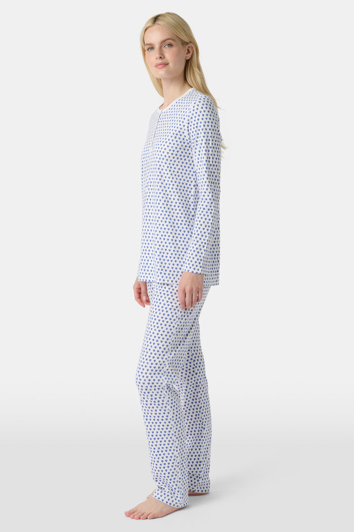 A person with light skin and long blonde hair stands barefoot in Roller Rabbit’s Hearts Pajamas—a matching set made from soft Pima cotton, featuring a white background with small blue and red heart patterns.