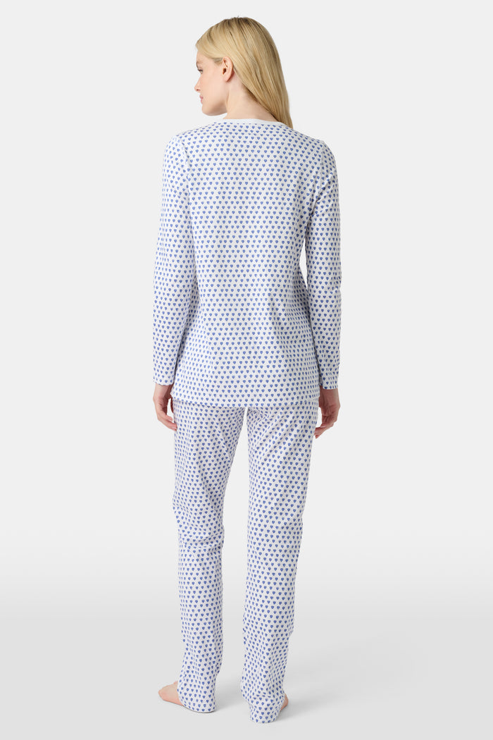 A person with light skin and long blonde hair stands barefoot in Roller Rabbit’s Hearts Pajamas—a matching set made from soft Pima cotton, featuring a white background with small blue and red heart patterns.