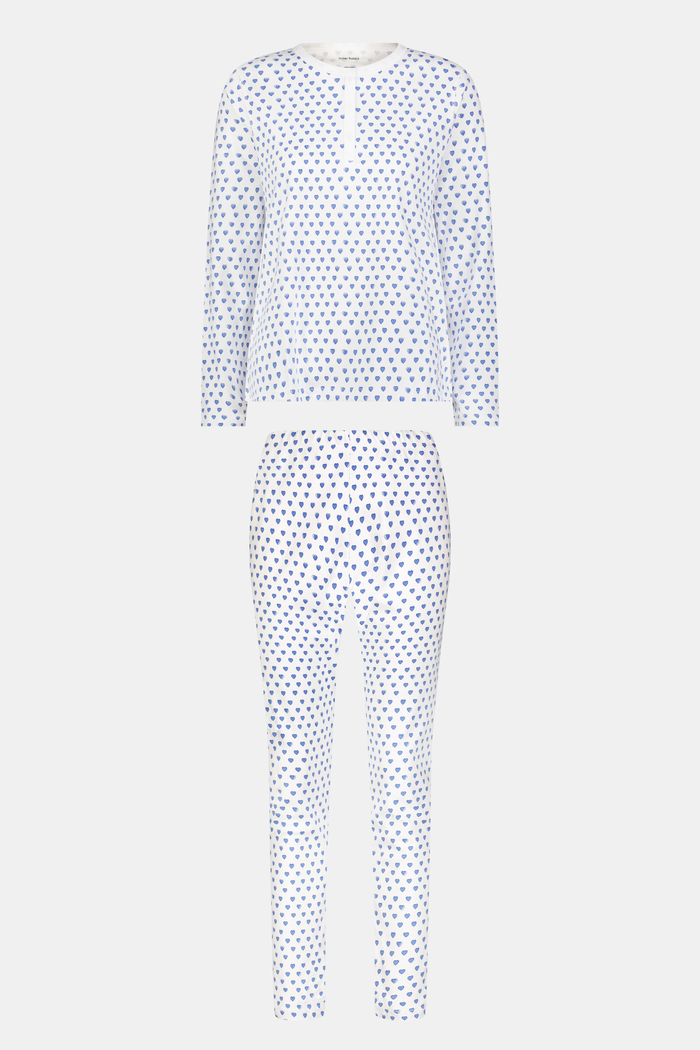 A person with light skin and long blonde hair stands barefoot in Roller Rabbit’s Hearts Pajamas—a matching set made from soft Pima cotton, featuring a white background with small blue and red heart patterns.