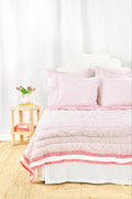 A neatly made bed features the Roller Rabbit Hearts Quilt in pink and white cotton voile with matching pillows, beside a small wooden table holding stacked books and a vase of pink tulips, set against a light background.