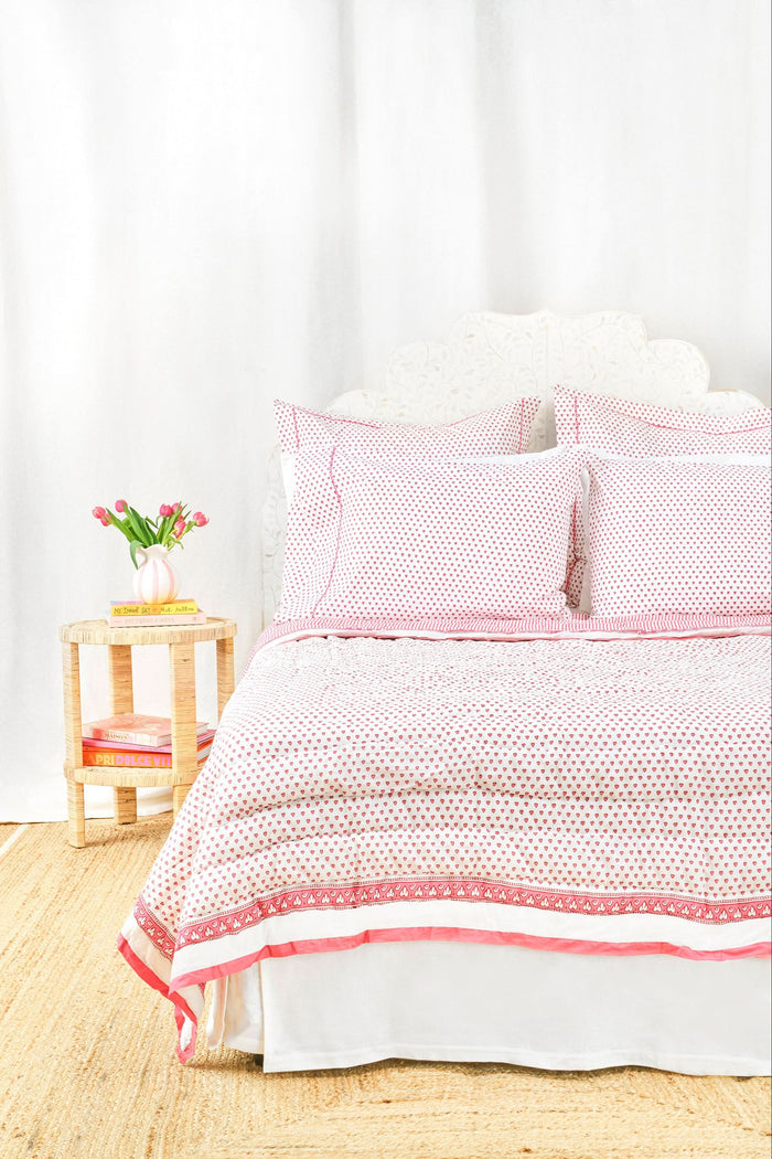 A neatly made bed features the Roller Rabbit Hearts Quilt in pink and white cotton voile with matching pillows, beside a small wooden table holding stacked books and a vase of pink tulips, set against a light background.