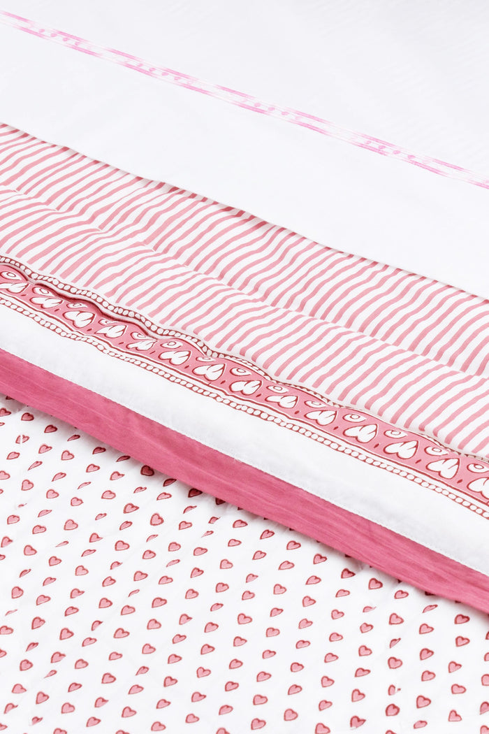 A neatly made bed features the Roller Rabbit Hearts Quilt in pink and white cotton voile with matching pillows, beside a small wooden table holding stacked books and a vase of pink tulips, set against a light background.