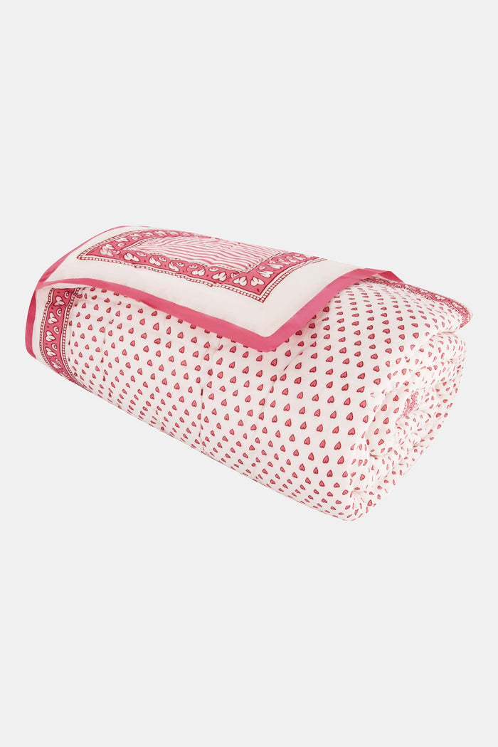 A neatly made bed features the Roller Rabbit Hearts Quilt in pink and white cotton voile with matching pillows, beside a small wooden table holding stacked books and a vase of pink tulips, set against a light background.