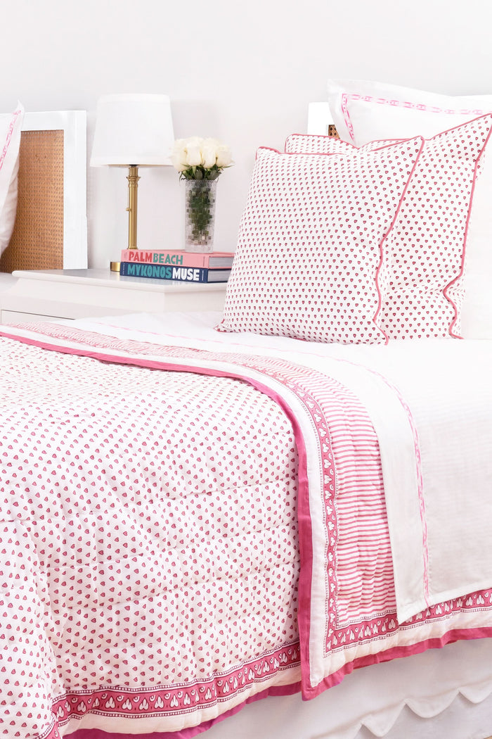 A neatly made bed features the Roller Rabbit Hearts Quilt in pink and white cotton voile with matching pillows, beside a small wooden table holding stacked books and a vase of pink tulips, set against a light background.