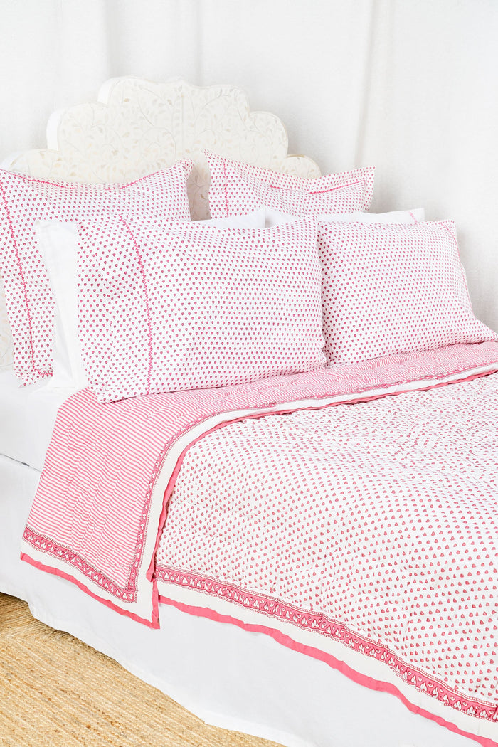 A neatly made bed features the Roller Rabbit Hearts Quilt in pink and white cotton voile with matching pillows, beside a small wooden table holding stacked books and a vase of pink tulips, set against a light background.