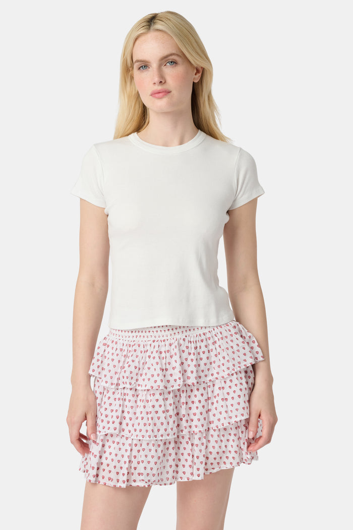A woman with long blonde hair wears a white short-sleeve t-shirt and the Roller Rabbit Hearts Siggy Skirt, a white tiered mini skirt with pink heart print, standing against a plain light background.