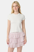 A woman with long blonde hair wears a white short-sleeve t-shirt and the Roller Rabbit Hearts Siggy Skirt, a white tiered mini skirt with pink heart print, standing against a plain light background.