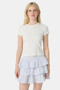 A woman with long blonde hair wears a fitted white short-sleeve t-shirt and the Roller Rabbit Hearts Siggy Skirt—a white ruffle mini skirt with a blue heart print—while standing against a plain white background.