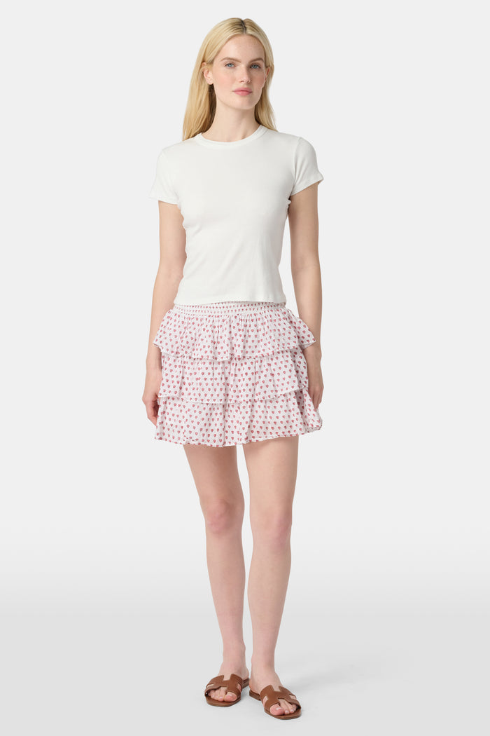 A woman with long blonde hair wears a white short-sleeve t-shirt and the Roller Rabbit Hearts Siggy Skirt, a white tiered mini skirt with pink heart print, standing against a plain light background.