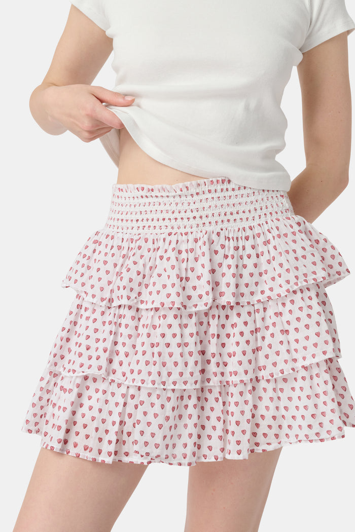 A woman with long blonde hair wears a white short-sleeve t-shirt and the Roller Rabbit Hearts Siggy Skirt, a white tiered mini skirt with pink heart print, standing against a plain light background.