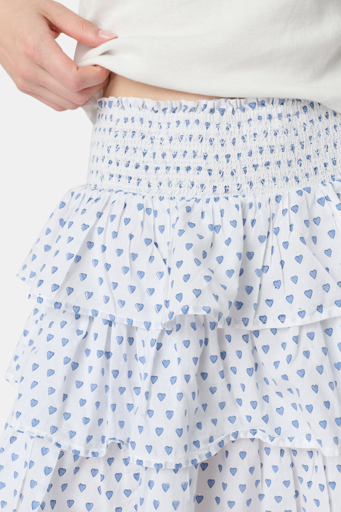 A woman with long blonde hair wears a fitted white short-sleeve t-shirt and the Roller Rabbit Hearts Siggy Skirt—a white ruffle mini skirt with a blue heart print—while standing against a plain white background.