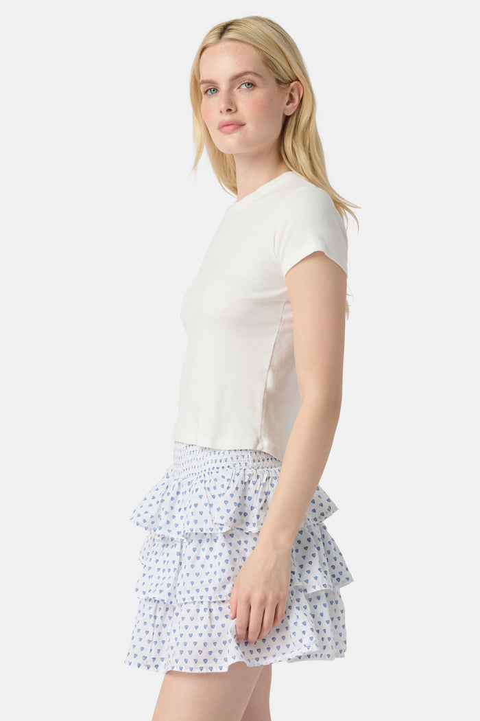 A woman with long blonde hair wears a fitted white short-sleeve t-shirt and the Roller Rabbit Hearts Siggy Skirt—a white ruffle mini skirt with a blue heart print—while standing against a plain white background.