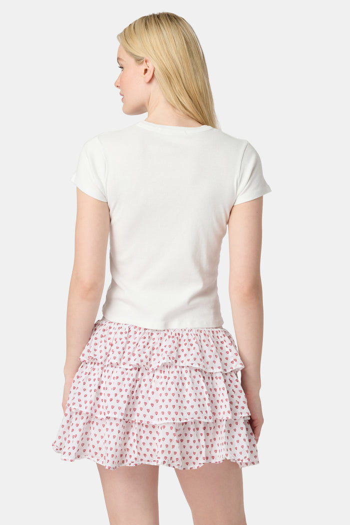 A woman with long blonde hair wears a white short-sleeve t-shirt and the Roller Rabbit Hearts Siggy Skirt, a white tiered mini skirt with pink heart print, standing against a plain light background.