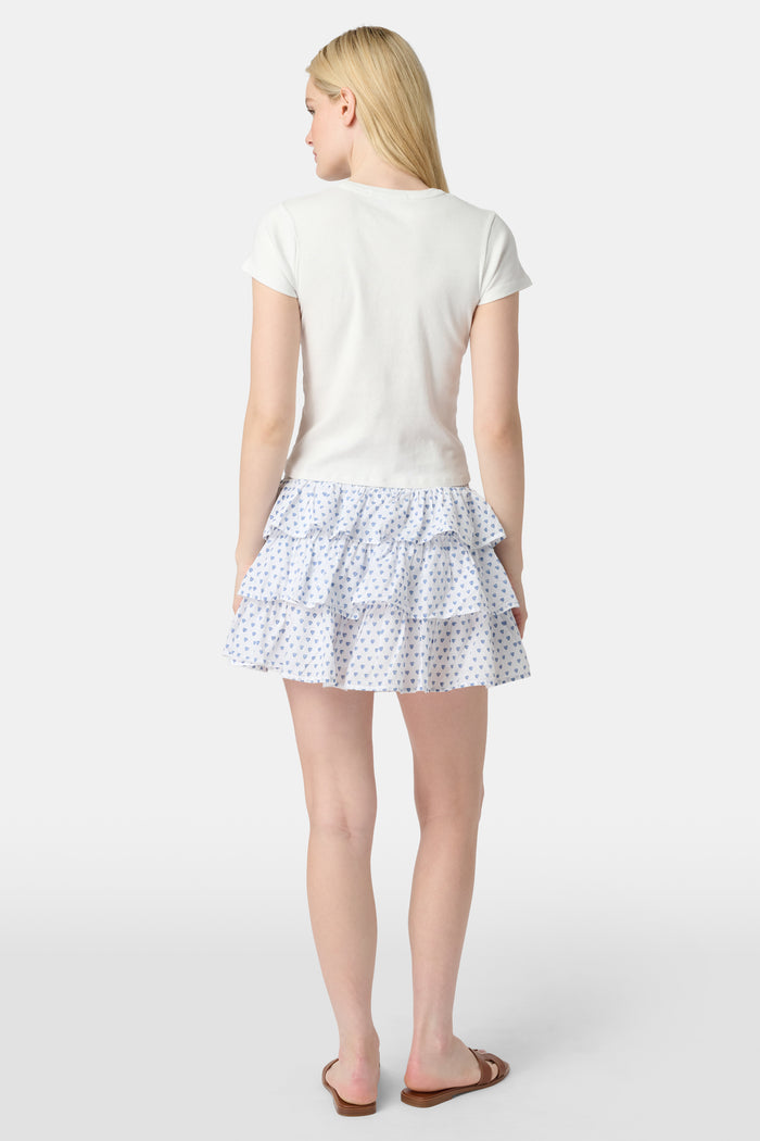 A woman with long blonde hair wears a fitted white short-sleeve t-shirt and the Roller Rabbit Hearts Siggy Skirt—a white ruffle mini skirt with a blue heart print—while standing against a plain white background.