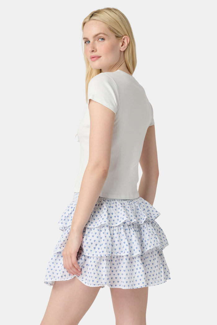 A woman with long blonde hair wears a fitted white short-sleeve t-shirt and the Roller Rabbit Hearts Siggy Skirt—a white ruffle mini skirt with a blue heart print—while standing against a plain white background.