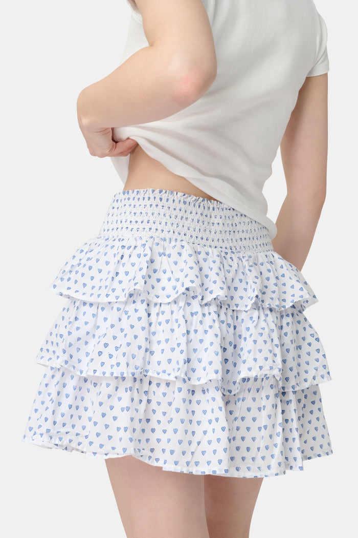 A woman with long blonde hair wears a fitted white short-sleeve t-shirt and the Roller Rabbit Hearts Siggy Skirt—a white ruffle mini skirt with a blue heart print—while standing against a plain white background.