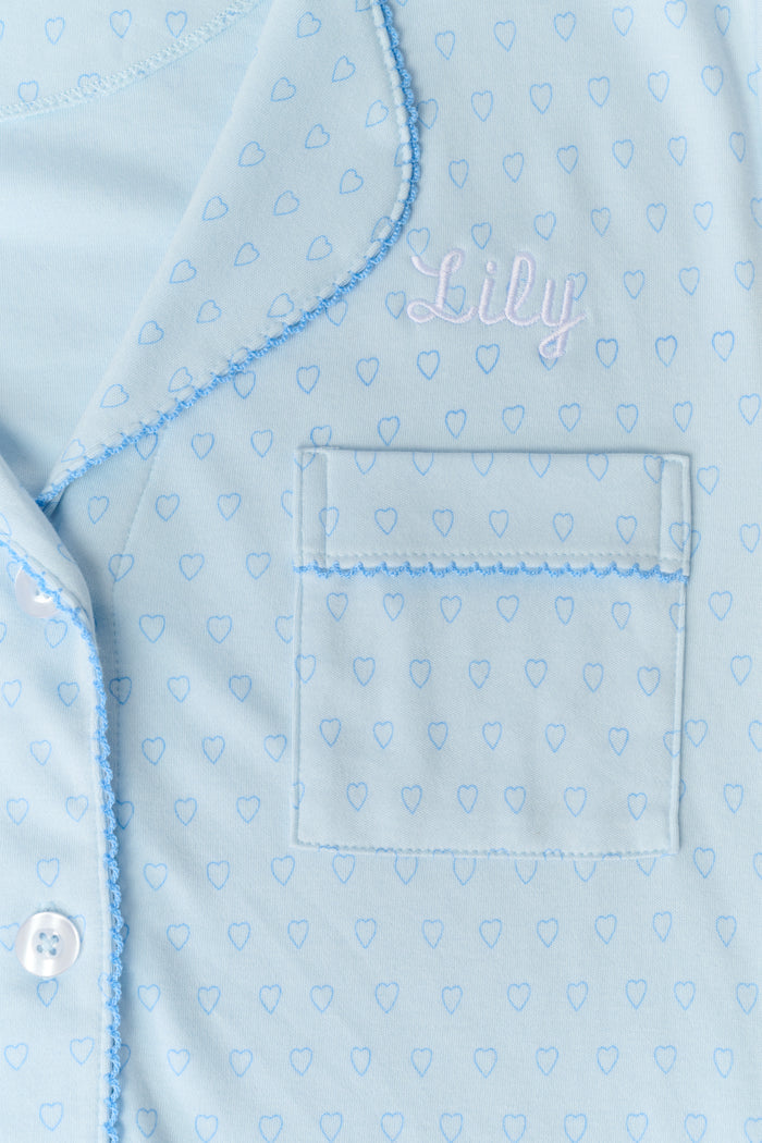 A woman wears Roller Rabbit's Hearts Tonal Wyatt Polo Pajamas—light blue, short-sleeved Pima cotton with a collar and chest pocket—paired with matching ruffled shorts, standing against a plain light background.
