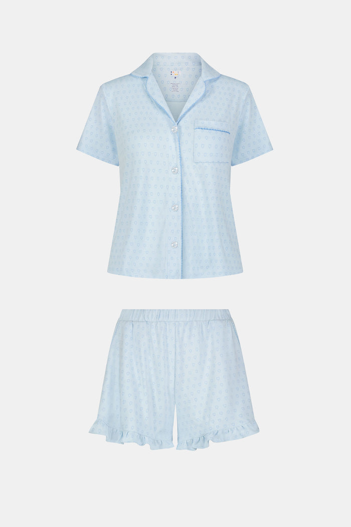 A woman wears Roller Rabbit's Hearts Tonal Wyatt Polo Pajamas—light blue, short-sleeved Pima cotton with a collar and chest pocket—paired with matching ruffled shorts, standing against a plain light background.