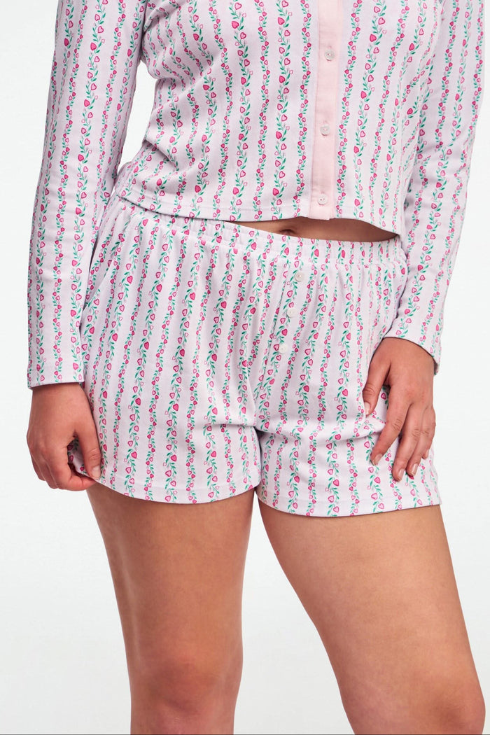 A person wears the Roller Rabbit Hearts Vine Brooklyn Boxer—matching long-sleeve, button-up pajama top and shorts with a pink, green, and white floral stripe pattern—standing against a white background.