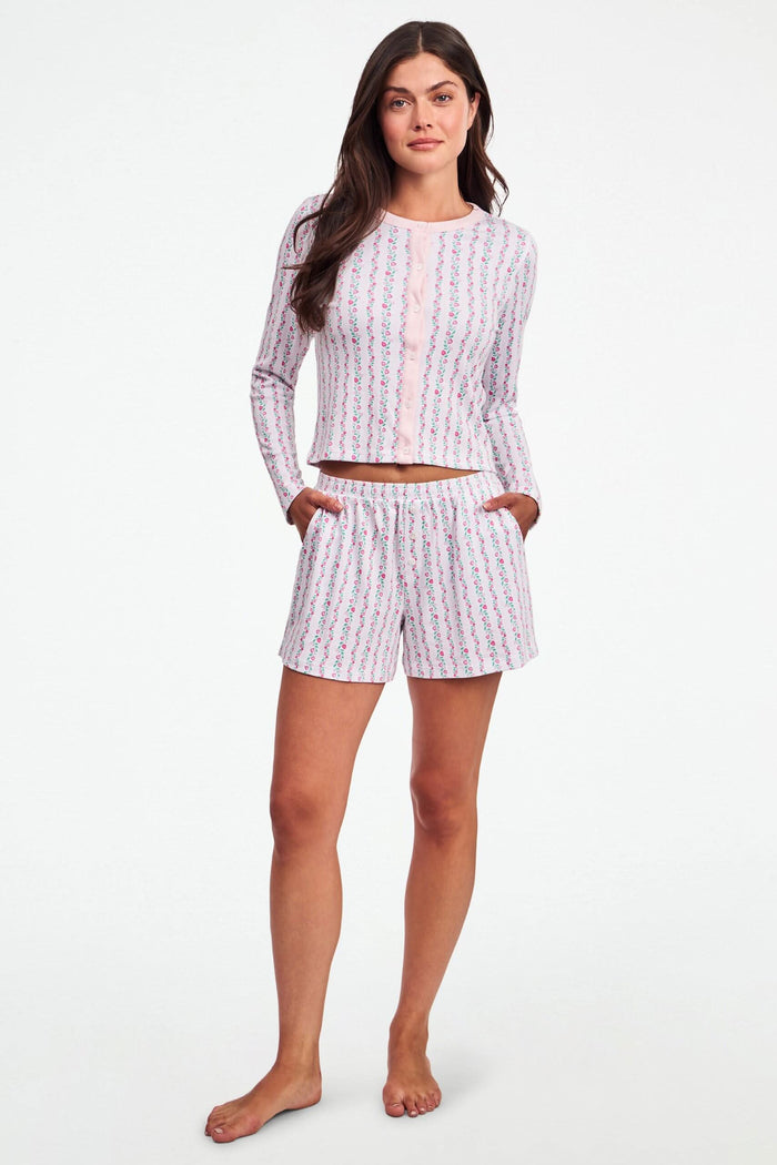 A person wears the Roller Rabbit Hearts Vine Brooklyn Boxer—matching long-sleeve, button-up pajama top and shorts with a pink, green, and white floral stripe pattern—standing against a white background.