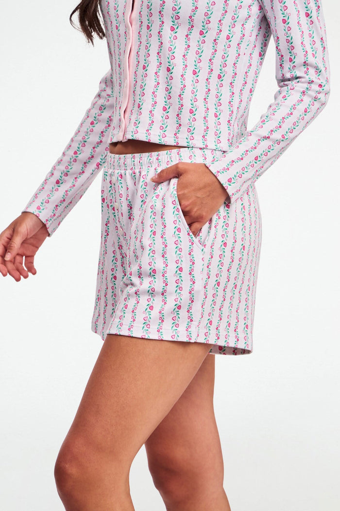 A person wears the Roller Rabbit Hearts Vine Brooklyn Boxer—matching long-sleeve, button-up pajama top and shorts with a pink, green, and white floral stripe pattern—standing against a white background.
