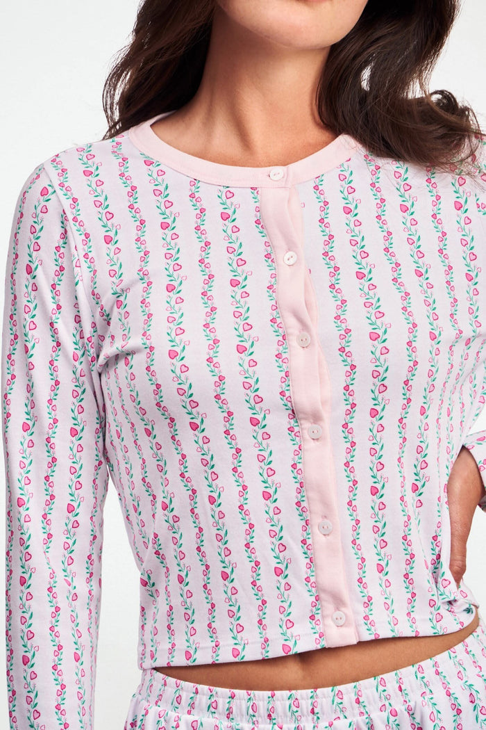 A woman wears the Roller Rabbit Hearts Vine Nessa Cardigan, a pink long-sleeve pima cotton sweater with small green and pink floral vine print and white front buttons. Her hand is on her hip, her face partially out of frame.