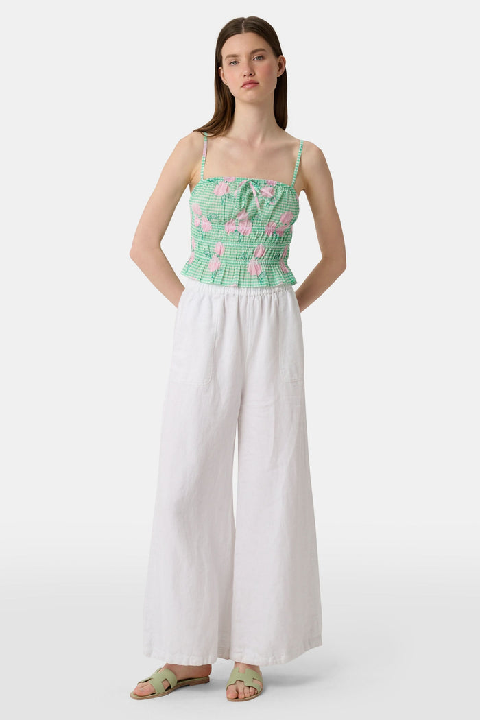 A woman wears the Roller Rabbit Hydrangea Gingham Aurelia Top—a green and white gingham crop top with pink hydrangea florals, thin straps, and a ruffled hem—paired with white pants against a plain white background.