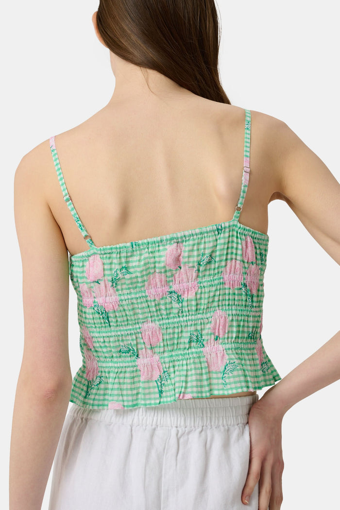 A woman wears the Roller Rabbit Hydrangea Gingham Aurelia Top—a green and white gingham crop top with pink hydrangea florals, thin straps, and a ruffled hem—paired with white pants against a plain white background.