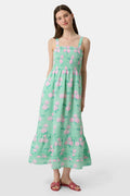 A woman wears the Roller Rabbit Hydrangea Gingham Bristol Dress, a long sleeveless style with pink florals, ruffled hem, and wide straps. She pairs it with pink sandals, standing with one hand by her side and the other on her leg.