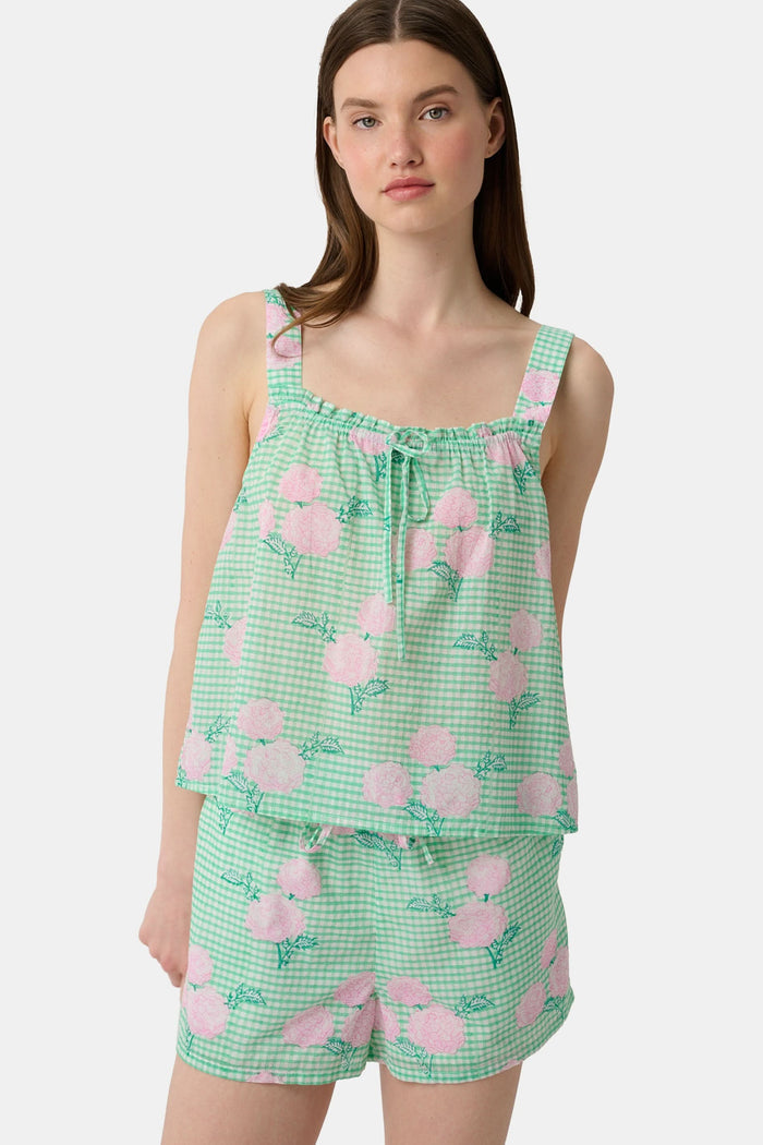 A woman with straight brown hair and a neutral expression wears the Roller Rabbit Hydrangea Gingham Cooper Set—green and white with pink floral prints. She stands with her hands in her shorts’ pockets and wears pink sandals.