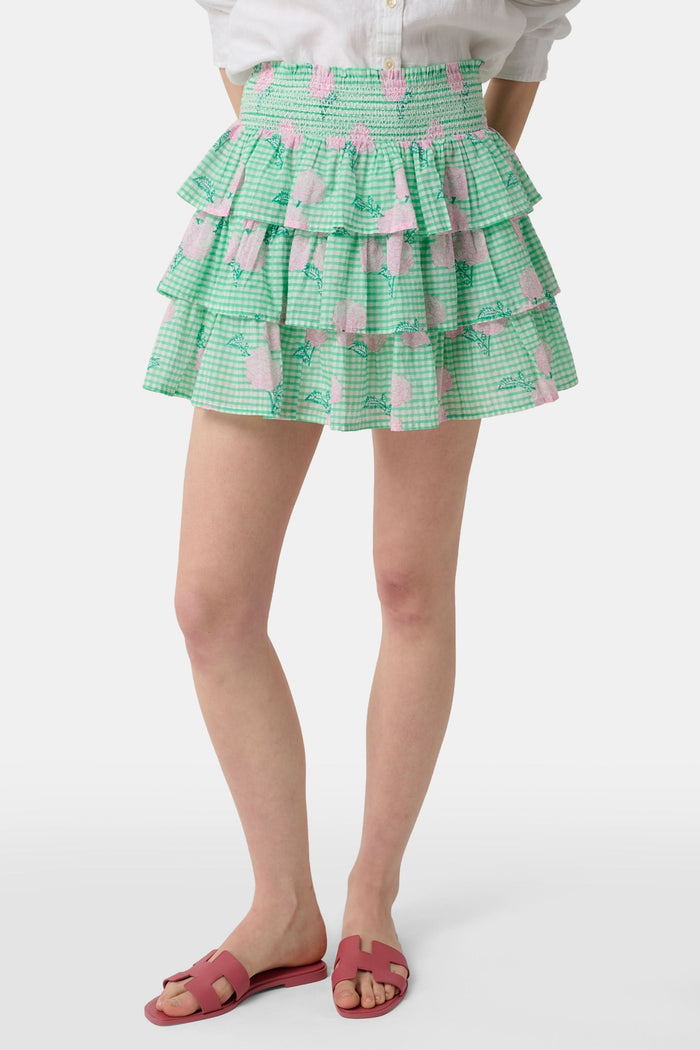 A person models the Roller Rabbit Hydrangea Gingham Siggy Skirt, featuring green and white checks with pink florals, paired with a tucked-in white shirt and red flat sandals. The image displays the skirt from waist to feet.