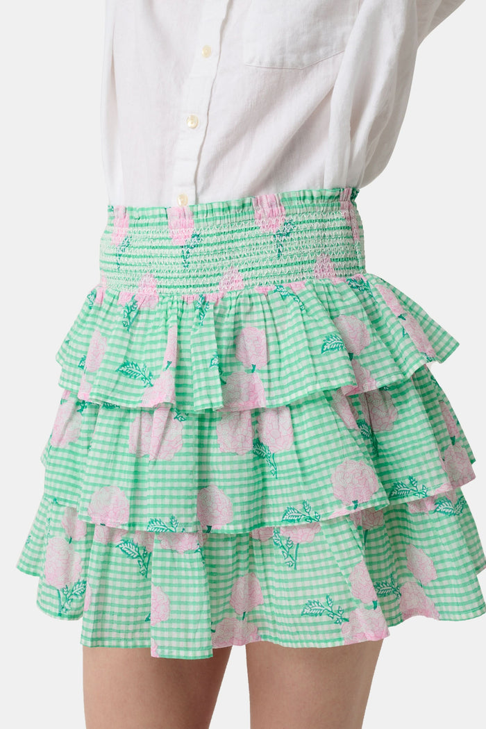 A person models the Roller Rabbit Hydrangea Gingham Siggy Skirt, featuring green and white checks with pink florals, paired with a tucked-in white shirt and red flat sandals. The image displays the skirt from waist to feet.