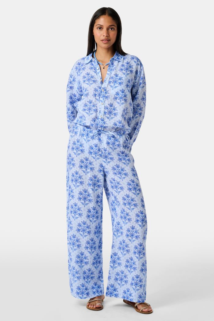 A woman stands against a plain light background wearing the Roller Rabbit Indigo Garden Boyfriend Shirt, a button-up long-sleeve pajama top in a blue floral print paired with matching wide-leg pants.
