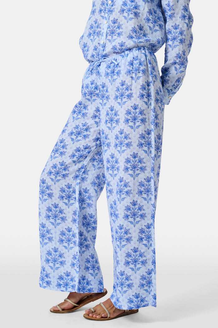 A woman stands against a plain light background wearing the Roller Rabbit Indigo Garden Boyfriend Shirt, a button-up long-sleeve pajama top in a blue floral print paired with matching wide-leg pants.