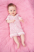 A baby with light skin and light brown hair lies on a pink and white striped blanket wearing the Roller Rabbit Infant Hearts Polo Onesie, looking slightly to the side.